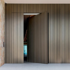 Seamless flush-mount pivot door integrated into matching deep bronze metallic wall cladding with vertical linear grooves.