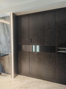 Flush-mount pivot armored door in deep matte grey concrete patina with precise geometric grid and brushed stainless horizontal mid-band, seamlessly integrated into marble wall in modern minimalist interior.
