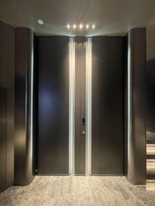A full-height, dark metallic armored double door with two vertical bright white LED light strips running from floor to ceiling, integrated smart lock, and curved metallic side pillars in a luxury marble interior.