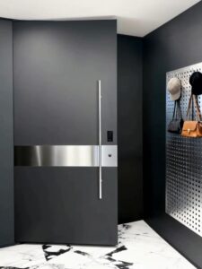 A tall, modern matte black armored door with a wide horizontal brushed stainless steel inlay, a slim floor-to-ceiling vertical silver handle, and an integrated smart lock, shown in a minimalist interior with marble flooring.