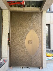 A tall, luxurious armored door made of hand-chiseled brass with a radiating sunburst pattern and a smooth almond-shaped central panel, installed in a light granite facade.