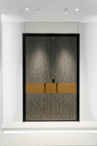 A tall double armored door with a grey fabric-like textured finish, featuring a wide horizontal brushed gold metal band in the center with integrated vertical pull handles and a smart lock.