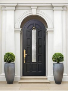 Classical arched single front door in midnight black, featuring a central narrow frosted glass panel, ornate gold handle and knocker, set against a white neoclassical stone facade with symmetrical topiaries.