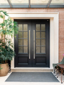 Modern black French-style double entrance door with 8-pane textured glass lites and long vertical stainless steel handles, set in a light stone architrave under a glass canopy.