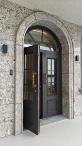 A heavy dark-colored armored entry door with rectangular glass window grids and ornate brass handles, partially open to reveal door thickness, installed within a gray granite arched stone wall.