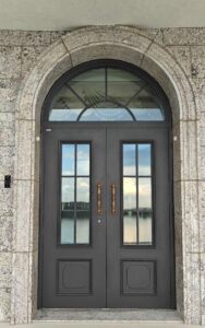 Full front view of a dark charcoal armored double door with rectangular glass grids and antique brass handles, set within a massive arched granite stone wall.