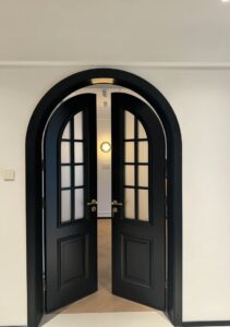 Full view of a modern matte black French-style double entrance door with long vertical stainless steel pull handles, installed on a minimalist light-colored porch.