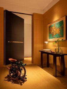 Flush-mount pivot door in ultra-matte jet black with single slim horizontal brushed gold pull, seamlessly integrated into matching dark wall cladding in luxury warm-lit interior corridor with art and folding bike.