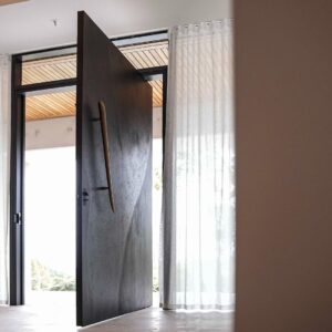 Flush-mount pivot door in deep matte charcoal with 3D carved organic dune wave relief texture and full-height sculptural walnut organic pull, seamlessly integrated into modern natural-light interior with sheer curtains.
