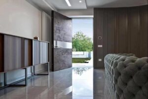 Flush-mount pivot door in matte concrete grey patina with broad brushed stainless mid-band, slim horizontal pull and integrated smart lock panel, seamlessly integrated into modern luxury interior with garden view and curved sofa.