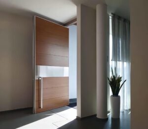 Flush-mount pivot door in natural walnut vertical grain with broad brushed stainless horizontal bands and full-height slim vertical stainless pull, seamlessly integrated into modern minimalist interior with natural light and cylindrical column.