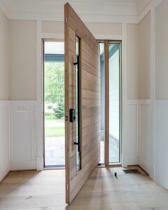 Flush-mount pivot door in light natural walnut horizontal grain with broad matte black horizontal bands, integrated slim vertical glass side panel, and full-height slim matte black vertical pull, seamlessly integrated into modern residential interior with abundant natural light and outdoor view.