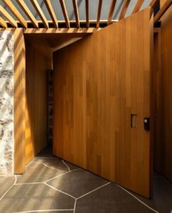 Luxury bespoke wood pivot door with vertical plank texture and a recessed square integrated handle in a modern home.