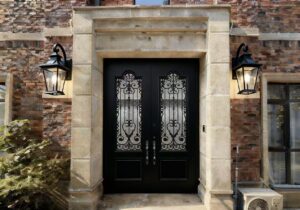 Full view of a matte black armored double door with ornate wrought iron scrolls and obscure glass, installed within a massive rectangular stone portal on a brick facade with traditional lanterns.