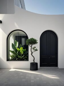 A minimalist white exterior wall featuring a large arched black armored door and a matching arched glass window reflecting tropical plants, with a Bonsai tree in between.