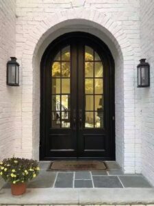 A symmetrical matte black arched double armored door with glass window grids, installed in a white brick recessed arched entryway with traditional black lanterns and a welcome mat.