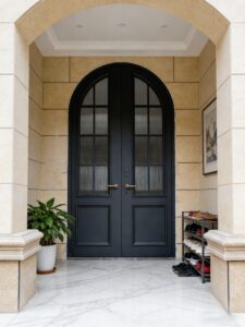 Classic French-style arched double pivot armored door in deep matte black with arched ribbed glass panels and gold hardware, set in light stone arched entrance with marble floor.