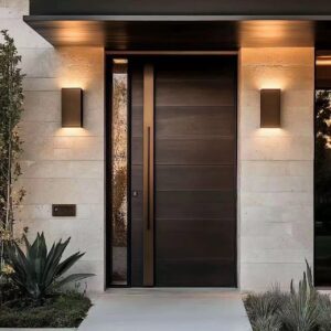 A modern single armored door with a dark wood-grain texture and a vertical brushed gold decorative strip, featuring a long black pull handle and a narrow vertical glass sidelight, installed in a light-toned stone wall with modern sconce lighting.