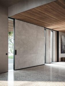 A massive, minimalist sliding armored door made of large-format grey sintered stone panels, featuring an ultra-slim black frame and a flush-integrated vertical handle in a modern interior with wood ceilings.