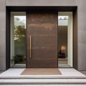 A massive weathered steel-textured single pivot door with a tall vertical bronze/gold handle, flanked by two large glass sidelights in a minimalist grey concrete architectural facade.