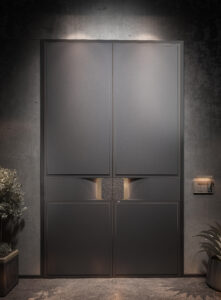A grand symmetric double armored door in charcoal grey with four recessed panels, thin gold/bronze outlines, and a central square sculptural element that glows with warm backlighting, set against a dark industrial concrete wall.