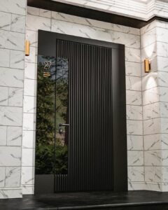 A tall, modern black armored door with vertical fluted ribbing and a left-side glossy black glass panel displaying the apartment number '27' in gold, installed in a white marble-clad hallway with gold wall sconces.