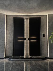 A grand symmetric double armored door in deep black wood grain, featuring a vertical glowing light strip in the center and two horizontal recessed light bars behind the handles, set against a grey concrete wall.