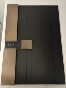 A tall, modern asymmetric armored door in matte black, featuring large rectangular textured bronze panels on the left, a central square bronze handle, and the gold number '888' integrated into the facade.