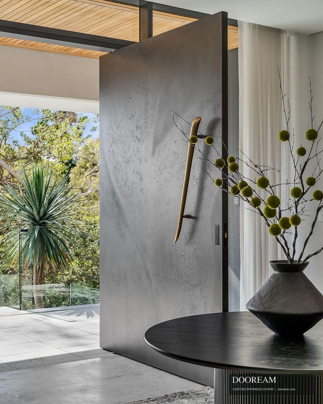 Design reference showing an open grey-toned pivot entrance with a sculptural warm-metal-tone pull in a contemporary interior